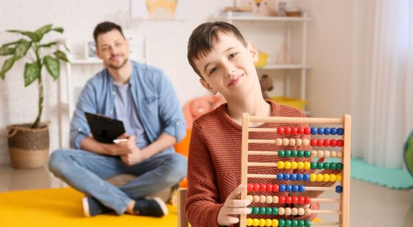 طفل يلعب بأداة عدّ ملونة بينما يجلس والد في الخلفية يحمل جهازًا لوحيًا. الغرفة مليئة بالألوان، مع نبات في الزاوية وسجاد أصفر.