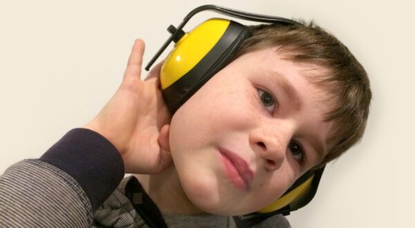 طفل يرتدي سماعات أذن صفراء وسوداء، يميل برأسه قليلاً إلى الجانب ويضع يده على أذنه، مع تعبير فضولي على وجهه.