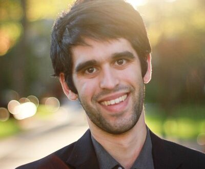 شاب مبتسم يرتدي بدلة داكنة، يقف في بيئة خارجية مضاءة بأشعة الشمس. خلفيته تحتوي على طبيعة مشمسة مع تأثير ضبابي.