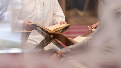 رجل يرتدي ثوبًا تقليديًا يحمل كتابًا مفتوحًا في مكان دراسي، مع وجود شخص آخر يجلس بجانبه، خلف ستارة شفافة.