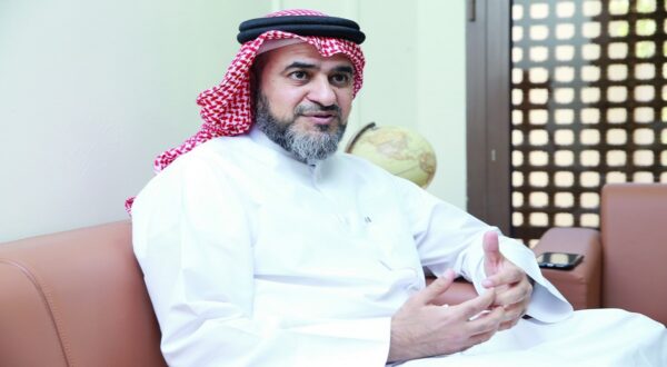 رجل يرتدي الزي التقليدي السعودي، يبتسم ويتحدث أثناء جلوسه على أريكة. تظهر في الخلفية خريطة، مما يدل على مكان مريح ومهني.