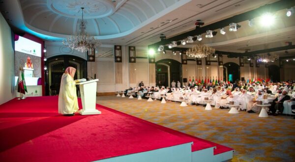 مؤتمر يتضمن متحدثًا على منصة أمام جمهور كبير في قاعة فسيحة، مع أعلام دول متعددة معروضة في الخلفية. المقاعد مرتبة بشكل جيد مع حضور رجال يرتدون الملابس التقليدية البيضاء. إضاءة مناسبة وسقف مزخرف يضيف لمسة من الفخامة.