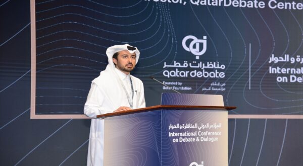 رجل يرتدي الزي التقليدي القطري يتحدث على منصة خلال مؤتمر دولي حول النقاش والحوار، مع وجود شعار مركز قطر للمناظرة في الخلفية.