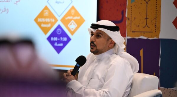 رجل يتحدث في حدث رسمي، يرتدي الزي التقليدي السعودي، ممسكًا بميكروفون. خلفه شاشة تعرض معلومات حول الفعالية. المكان مزين بألوان زاهية.