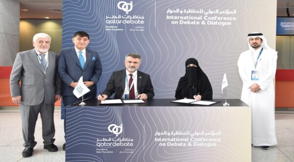 مؤتمر دولي حول المناظرة والحوار في قطر، يتضمن خمسة أشخاص يقفون حول طاولة توقيع. يظهر في الصورة رجلان وامرأتان ورجل آخر، حيث يقوم أحد الرجال بتوقيع وثيقة. الخلفية تظهر شعارات المؤتمر.