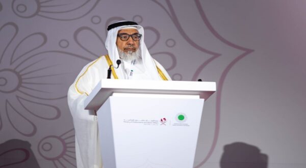 رجل مسن يرتدي الثوب العربي والعباءة، يتحدث من منصة مؤتمرات مع ميكروفون. خلفية بسيطة بتصميم زهري، تعكس حدثًا رسميًا أو مؤتمرًا.