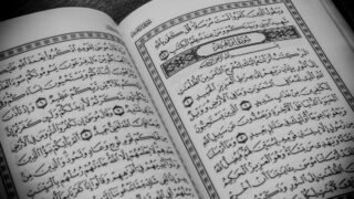 Open pages of an Arabic book with intricate calligraphy and decorative borders, featuring religious text. The book appears to be a Quran, emphasizing its cultural and spiritual significance.