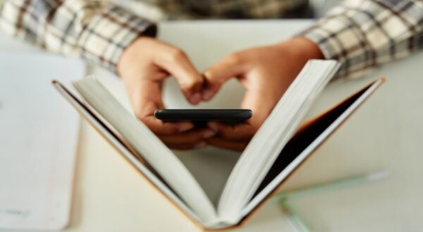 شخص يحمل هاتفًا محمولًا بيد واحدة بينما يقرأ كتابًا مفتوحًا بيد أخرى، يظهر مشهد يجمع بين التكنولوجيا والقراءة.