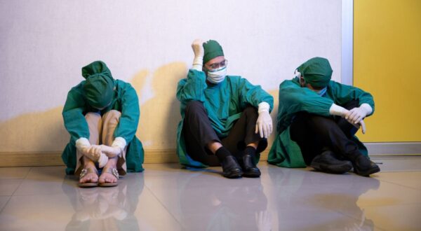 أطباء يرتدون ملابس جراحة خضراء وقناع وجه يجلسون على الأرض في ممر، يظهر عليهم التعب والقلق. يعكس المشهد الضغط النفسي الذي يواجهه الطاقم الطبي في ظل الظروف الصعبة.