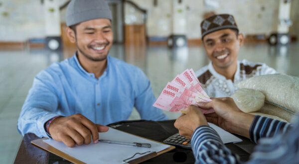 رجلان مبتسمان يتبادلان النقود داخل مسجد، يظهر أحدهما وهو يسلم أوراق نقدية لشخص آخر يجلس على طاولة. في الخلفية، تظهر أجواء المسجد مع تفاصيل معمارية هادئة.