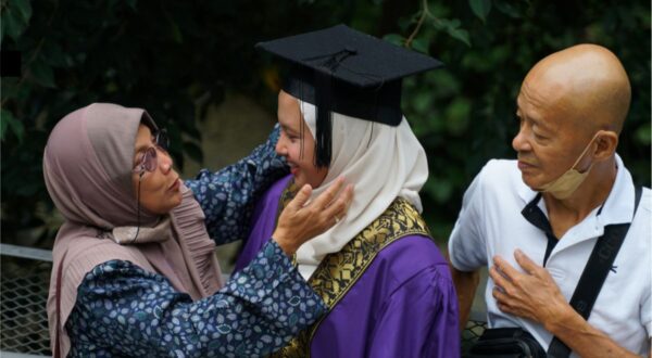 امرأة محجبة ترتدي قبعة تخرج وقميصًا بنفسجي، تتلقى التهاني من والدتها ووالدها في احتفال التخرج. الخلفية عشبية، مع greenery حولهم. تعبيرات الفرح والفخر واضحة على وجوههم.