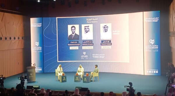 جلسة حوارية في مؤتمر دولي، حيث يتحدث ثلاثة متحدثين على المسرح أمام جمهور. الخلفية تحتوي على شعار المؤتمر وتصميم عصري. يركز النقاش على قضايا اقتصادية أو استراتيجية.