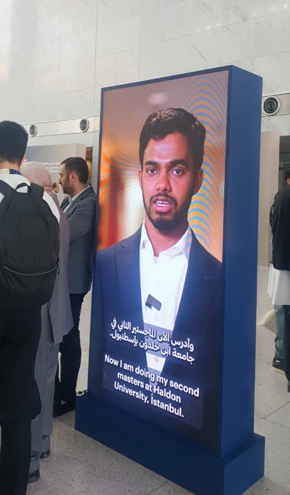 شاشة عرض رقمية تعرض شخصًا يتحدث عن دراسته في جامعة هالدون في إسطنبول، مع ترجمة للحديث. الخلفية تحتوي على مجموعة من الأشخاص يتجمعون حول الشاشة.