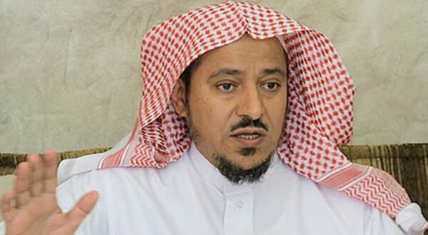 رجل يرتدي الزي التقليدي السعودي، بما في ذلك الشماغ والعقال، يتحدث بجدية. تظهر ملامح وجهه التركيز والانتباه، في خلفية بسيطة.