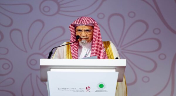 رجل مسن يرتدي لباسًا تقليديًا سعوديًا مع عقال، يتحدث من منصة في حدث رسمي. خلفية الصورة مزخرفة بألوان ناعمة، مع شعار وزارة الشؤون البلدية والقروية في أسفل المنصة.