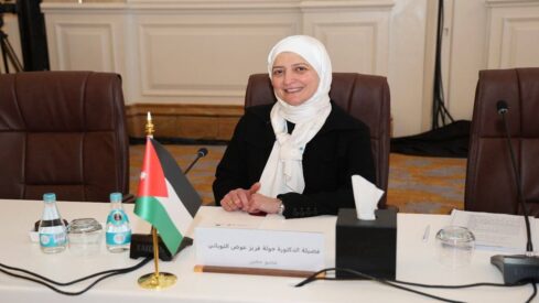 امرأة ترتدي الحجاب الأبيض تجلس على طاولة مؤتمر، مبتسمة، مع علمي الأردن وفلسطين أمامها، وزجاجات مياه وعلبة مناديل خلفها.