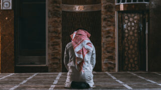 رجل يرتدي الزي التقليدي الإسلامي، يجلس متوجهاً نحو القبلة في مسجد، مع التركيز على الأجواء الروحية والهدوء الذي يحيط به.