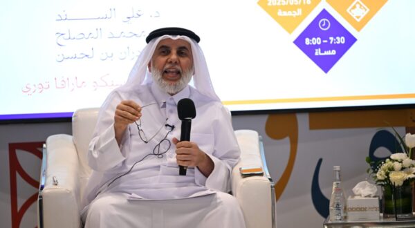 رجل يرتدي الزي التقليدي يتحدث في حدث، يحمل نظاراته بيده. خلفه شاشة تعرض معلومات عن الفعالية، تشمل التاريخ والوقت.
