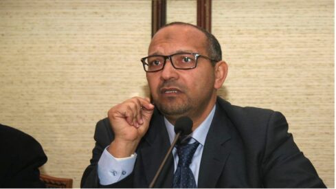 رجل يرتدي نظارات وقميصا رسميا، يتحدث أثناء مؤتمر أو اجتماع، مع تعبير وجه جاد. يظهر خلفه جدار بسيط.