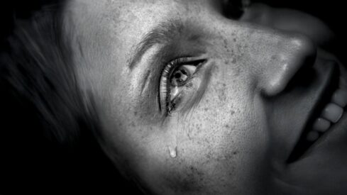 صورة مقربة لوجه امرأة مبتسمة مع دمعة تبرز من عينها، تظهر تفاصيل واضحة للبشرة والنمش. تعكس الصورة مشاعر مختلطة من الفرح والحزن، مع تباين قوي بين الظل والضوء، مما يضيف عمقًا عاطفيًا للمشهد.