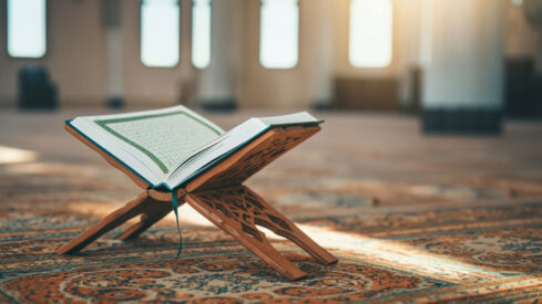 كتاب مفتوح على حامل خشبي داخل مسجد، مع سجادة مزخرفة وخلفية مضاءة بأشعة الشمس.