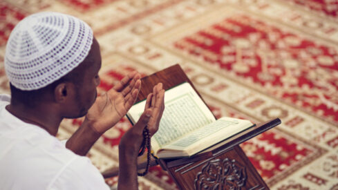 Muslim man in traditional attire praying while holding prayer beads, with an open Quran on a wooden stand, set on a patterned red carpet.