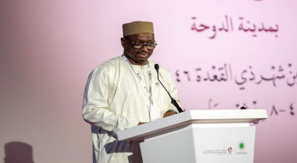 رجل يرتدي زيًا تقليديًا يتحدث من منصة في مؤتمر، مع خلفية وردية تحمل نصوصًا عربية.