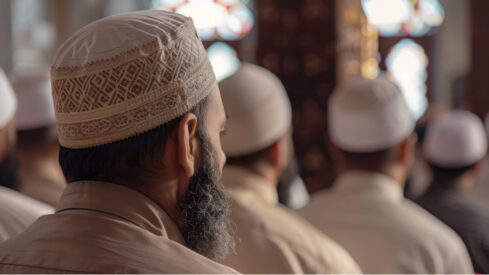 A group of men with traditional white caps sit in a mosque, focusing on a religious service. The scene highlights the cultural significance of communal worship in Islam, with intricate stained glass windows softly illuminating the interior.