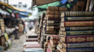 كتب قديمة موضوعة في أكوام مرتبة في سوق مكتظ، مع بائعين وزبائن في الخلفية. تبرز الأغلفة الملونة والتفاصيل الزخرفية للكتب، مما يُظهر التراث الثقافي والاهتمام بالقراءة.