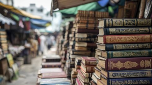 كتب قديمة موضوعة في أكوام مرتبة في سوق مكتظ، مع بائعين وزبائن في الخلفية. تبرز الأغلفة الملونة والتفاصيل الزخرفية للكتب، مما يُظهر التراث الثقافي والاهتمام بالقراءة.