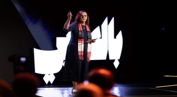 امرأة تتحدث على منصة، تحمل جهاز لوحي في يدها، وتوجه إشارات بيدها الأخرى. تتواجد خلفها خلفية مضاءة بكلمات عربية كبيرة، بينما يجلس الجمهور في الظل.