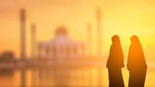 Two women in traditional attire stand silhouetted against a vibrant sunset, with an ornate mosque in the background. The scene captures a serene moment, highlighting cultural beauty and architectural elegance.