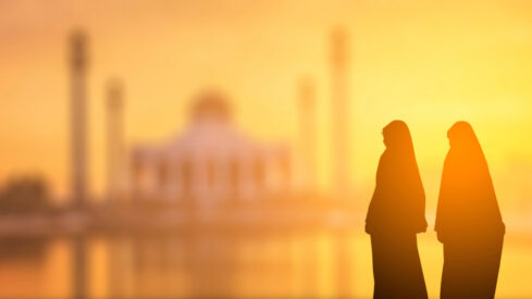 Two women in traditional attire stand silhouetted against a vibrant sunset, with an ornate mosque in the background. The scene captures a serene moment, highlighting cultural beauty and architectural elegance.