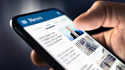 يد تظهر إصبعًا يتصفح تطبيق الأخبار على هاتف ذكي، مع عرض عناوين الأخبار الحديثة والصور.