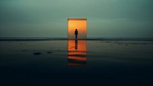 شخص يقف داخل إطار شفاف ينعكس على سطح مائي في مشهد غامض، مع ألوان دافئة في الأفق تعكس جمال الطبيعة.