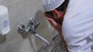 A man in a white kufi washes his face at a faucet in a bathroom, demonstrating a ritual cleansing. Water flows from the faucet, highlighting the importance of cleanliness in personal hygiene.