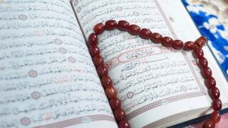 كتاب قرآن مفتوح مع نصوص عربية واضحة، ومسبحة حمراء على شكل قلب موضوعة فوقه، تعكس روحانية وعبادة.