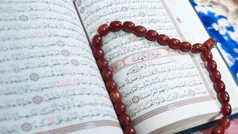 كتاب قرآن مفتوح مع نصوص عربية واضحة، ومسبحة حمراء على شكل قلب موضوعة فوقه، تعكس روحانية وعبادة.