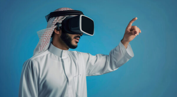 رجل يرتدي نظارات واقع افتراضي، يرتدي الزي التقليدي السعودي، ويشير بإصبعه نحو الأمام. خلفية زرقاء تعزز تجربة التكنولوجيا الحديثة.