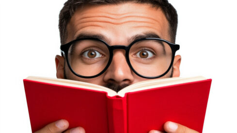 رجل يرتدي نظارات سميكة يحمل كتابًا أحمر، ينظر إلى الكاميرا بعينيه الواسعتين في تعبير عن الدهشة. يبرز المشهد شغف القراءة والاستكشاف.