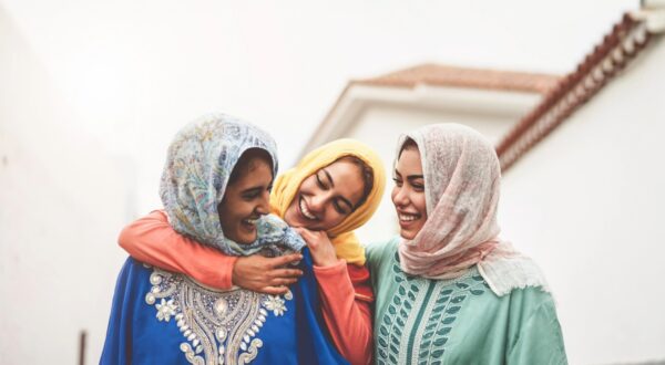 ثلاث نساء يرتدين أزياء تقليدية ملونة، يضحكن معًا في شارع هادئ. تظهر الألوان الزاهية للأوشحة والملابس، مما يعكس ثقافة غنية. الأجواء مبهجة وتعبر عن الصداقة والترابط بينهن.