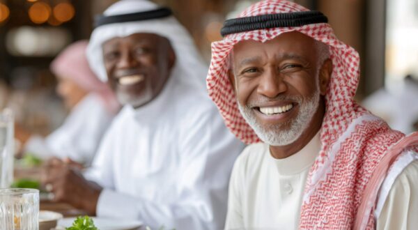 رجلان مسنّان يجلسان في مطعم، يرتدي أحدهما الزي التقليدي العربي مع شماغ، ويبتسمان أثناء تناول الطعام. تتواجد أطباق متنوعة على الطاولة، مما يبرز جوًا من الألفة والاحتفال.