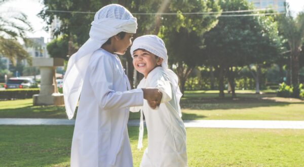 صبيان يتسابقان في حديقة، يرتديان ملابس تقليدية، يضحكان ويمسكان بأيدي بعضهما. الأشجار الخضراء والخلفية مشمسة، تعكس أجواء من الفرح والبراءة.