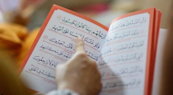 كتاب مفتوح يحتوي على نصوص باللغة العربية، مع التركيز على آيات قرآنية. تظهر يد تشير إلى إحدى الآيات. الخلفية ضبابية، مما يبرز تفاصيل الكتاب.