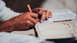 يد تظهر شخصًا يكتب في دفتر ملاحظات مع ورقة أخرى بجانبه، محاطًا بملاحظات مكتوبة وأدوات كتابة، تعكس عملية التفكير والتدوين.