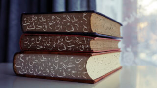 كتب ملونة موضوعة فوق بعضها، تحتوي على نصوص عربية مزخرفة. الخلفية تظهر نافذة مع ستائر شفافة، مما يضيف جواً هادئاً للمشهد.
