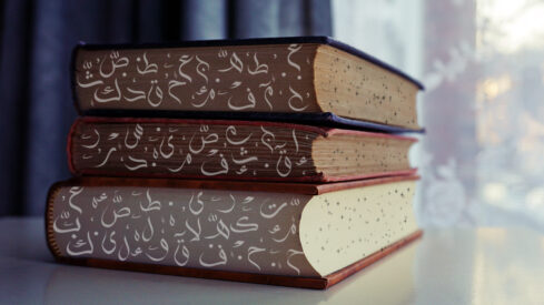 كتب ملونة موضوعة فوق بعضها، تحتوي على نصوص عربية مزخرفة. الخلفية تظهر نافذة مع ستائر شفافة، مما يضيف جواً هادئاً للمشهد.