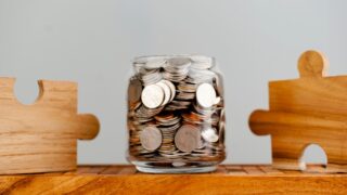 جرة زجاجية مملوءة بالعملات المعدنية موضوعة بين قطعتين خشبيتين على شكل أحجية، تعبر عن مفهوم الادخار والاستثمار.