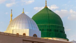 قبة المسجد النبوي في المدينة المنورة، تظهر قبتان مميزتان، واحدة فضية والأخرى خضراء، تحت سماء زرقاء مع سحب بيضاء. القبة الخضراء تعود لمسجد النبي محمد، وتعتبر رمزًا هامًا في الثقافة الإسلامية.