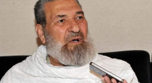 رجل مسن ذو لحية بيضاء يرتدي الإحرام، يجلس على أريكة ويتحدث مع شخص يحمل جهاز تسجيل. تعكس تعابير وجهه الجدية والاهتمام.